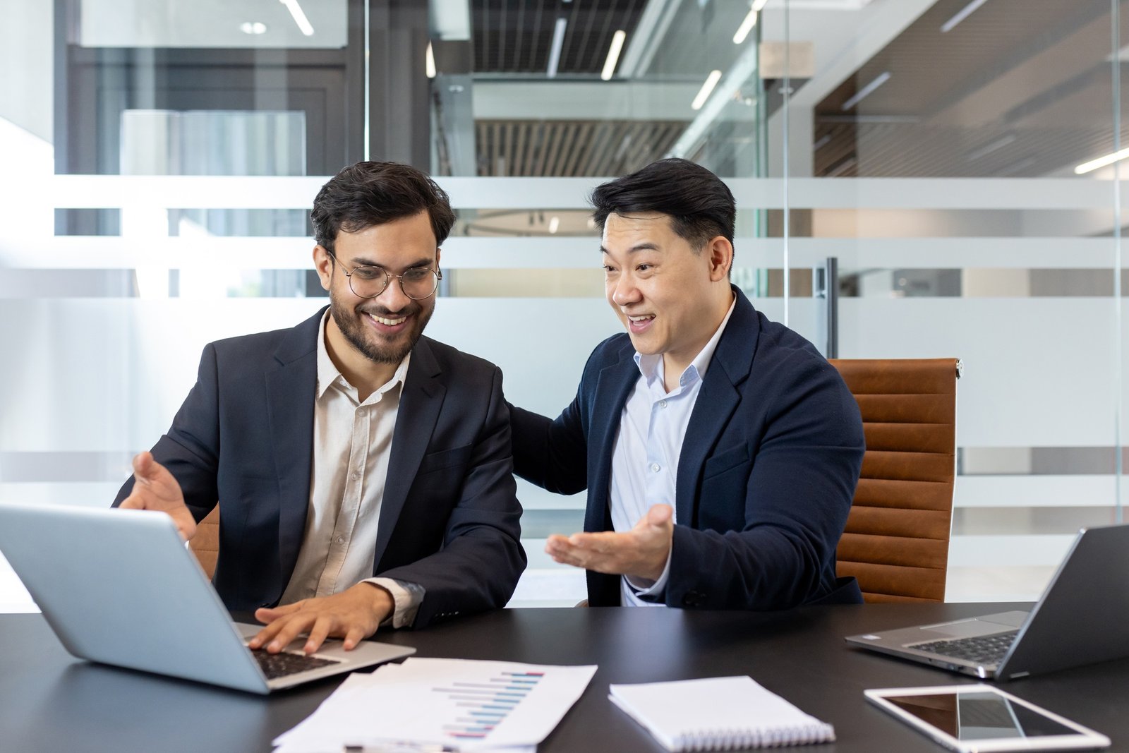 Male mentor at workplace helping intern learn new work project, two men in business suits working